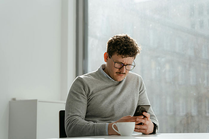 Man with glasses in a gray sweater, using phone by a window, caring attention.
