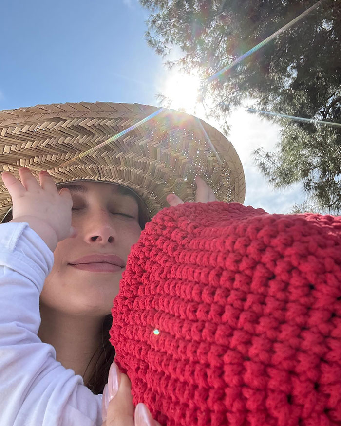 Woman in sun hat holding baby with red crochet blanket by the pool, sunlight streaming through trees. Keywords: Internet debate.