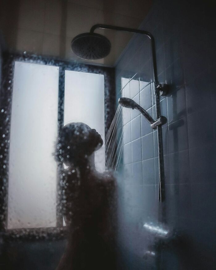 Silhouetted person under rainfall showerhead, water droplets on glass; exemplifying impractical elegance in a home setting.