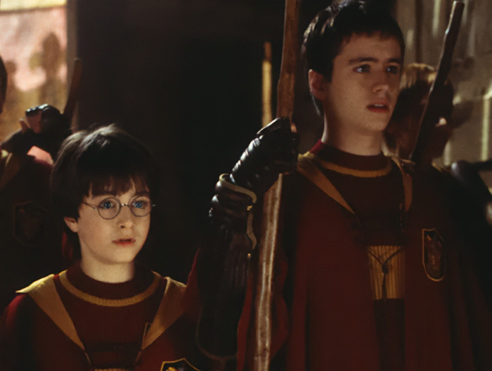Harry Potter star in red Quidditch gear with teammates, holding brooms in a castle corridor setting.