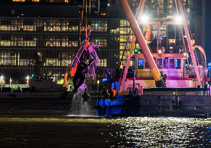 Rescue operation lifting crashed helicopter from New York water at night, against a background of city lights.