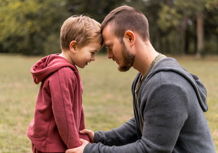 A man and child in hoodies touch foreheads affectionately in a park setting.