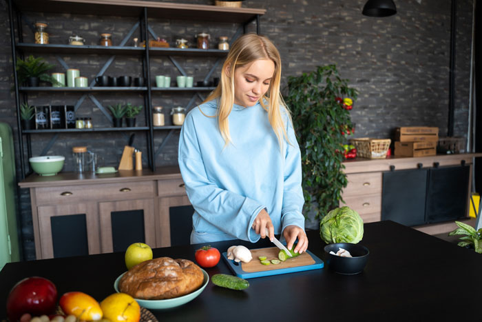 Woman in blue sweatshirt slicing cucumber, standing in a modern kitchen with various vegetables on the counter.
