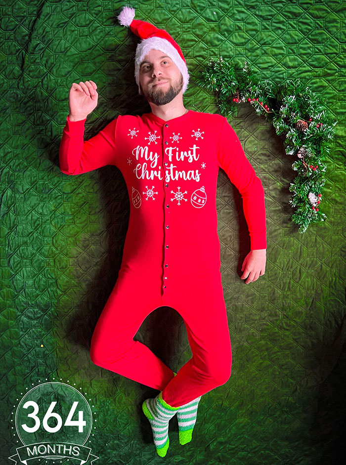 Man humorously dressed in a red onesie and Santa hat, lying on green background, ensuring life is never boring.