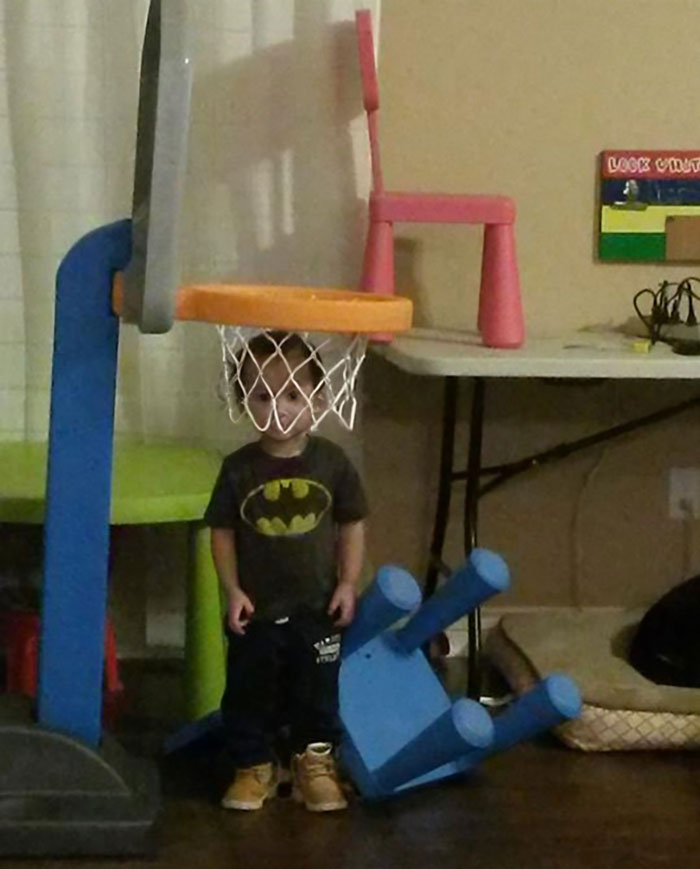 A child stuck in a toy basketball hoop with fallen chairs around, having a worse day than most.