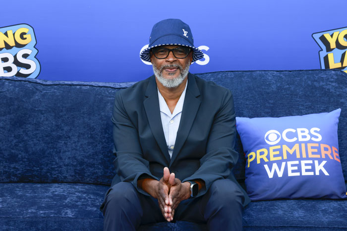 Film legend at CBS Premiere Week event, wearing a bucket hat and glasses, seated on a blue couch.