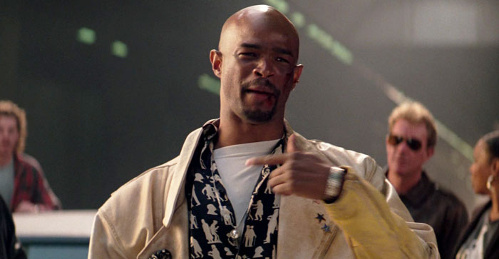 Film legend in a casual scene, wearing a beige jacket and patterned shirt, gesturing with his hand, smiling slightly.