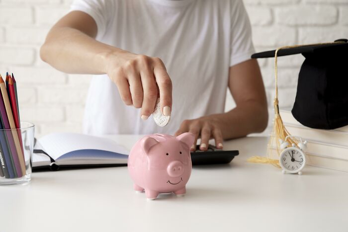 Person saving coin in piggy bank for first home tips, sitting at table with notebook and clock.