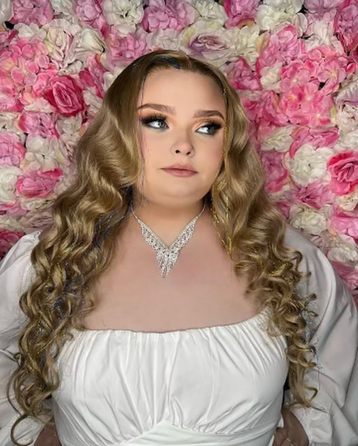 Young woman with long, curly hair in front of a floral backdrop, wearing a white dress and necklace.