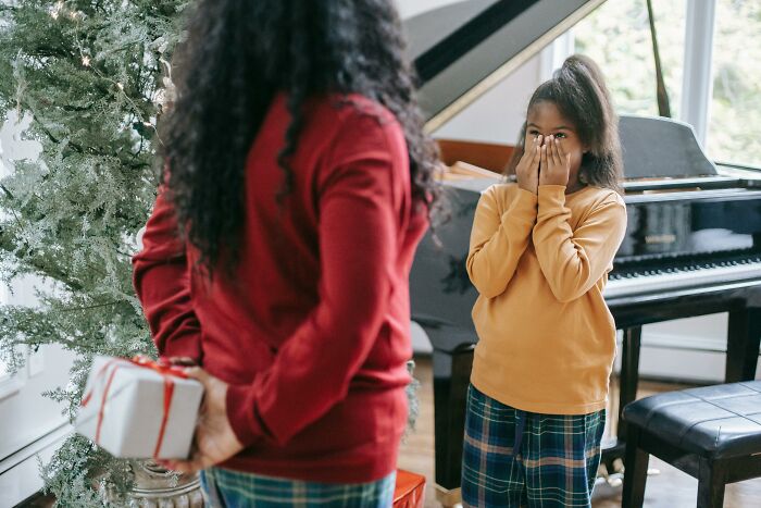 Child surprised by gift held behind adult, next to a piano and Christmas tree, expressing an unpopular opinion moment.