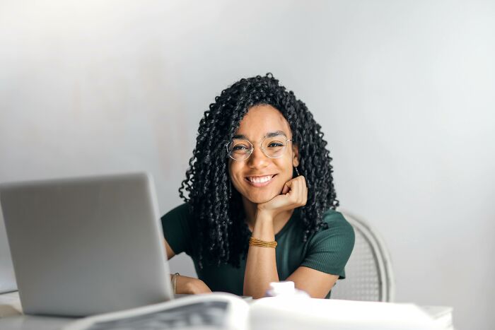 Smiling woman with glasses at a desk, pondering unpopular opinions, with a laptop and open book nearby.