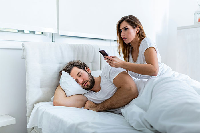 Woman in bed secretly checking sleeping partner's phone for cheating evidence.