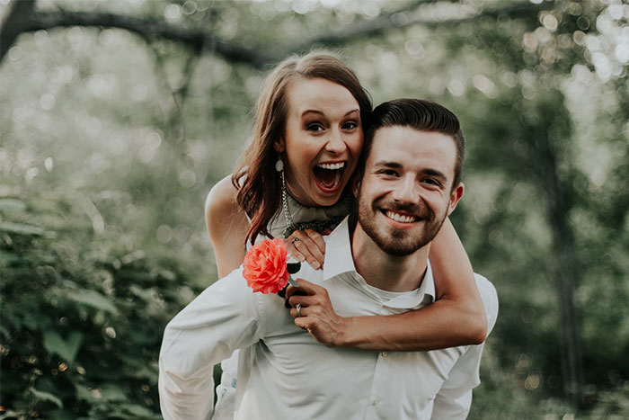 Smiling couple in a joyful embrace outdoors, holding a flower, amidst trees.
