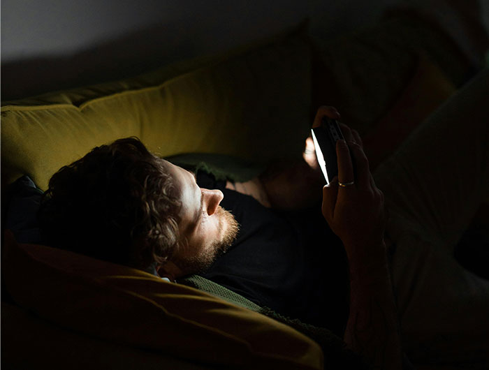 Man on couch in dark, illuminated by phone screen, possibly discovering partner's cheating.