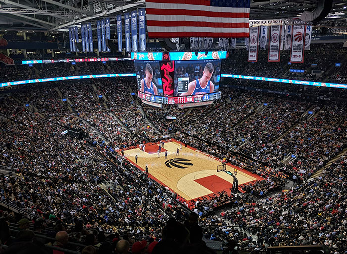 Packed basketball arena during an intense game with a large screen showing close-up of players.