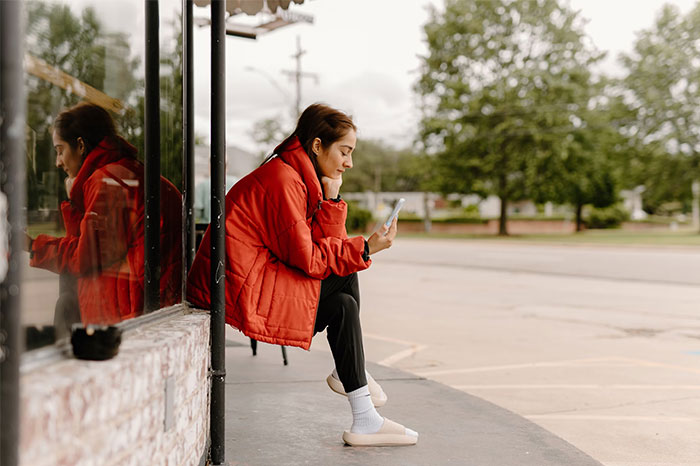 Person in red jacket looking at phone while sitting outside on a bench, contemplating a cheating partner.
