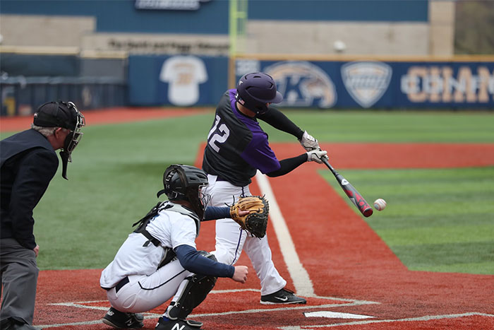 Baseball player in purple jersey swinging at the ball, with a catcher and umpire on the field.