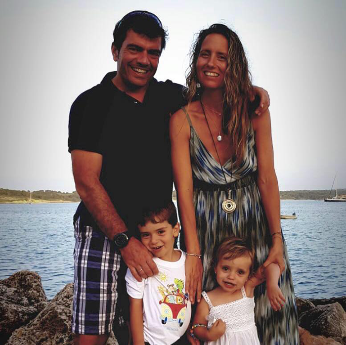 A smiling family of four stands outdoors near the water, with a sailboat visible in the background.