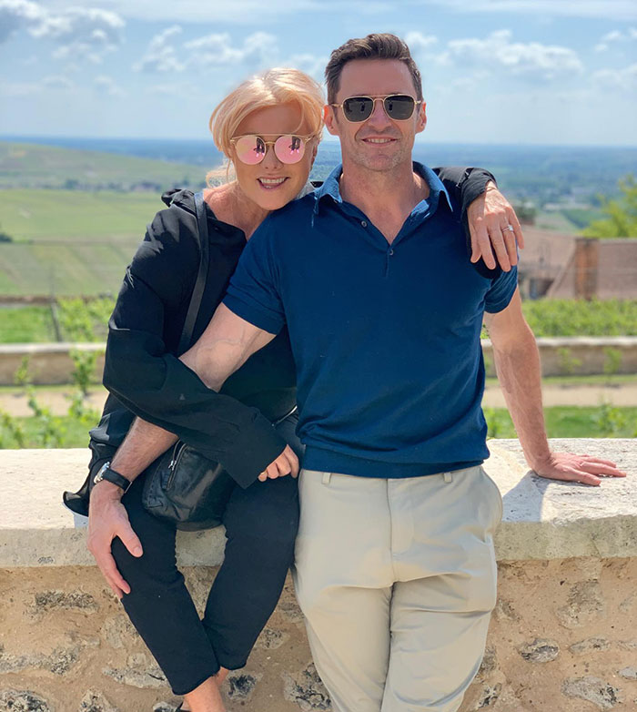 Hugh Jackman outdoors in sunglasses and blue polo, posing with a woman against a scenic background.