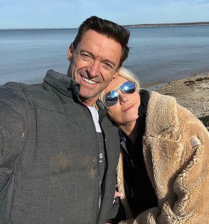 Man in a jacket taking a selfie with a woman in sunglasses by the water, smiling and enjoying a sunny day.