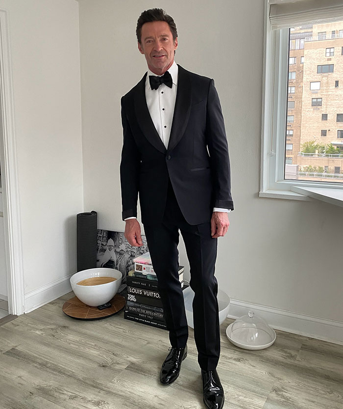 Man in a black tuxedo standing in a modern room, looking at the camera.