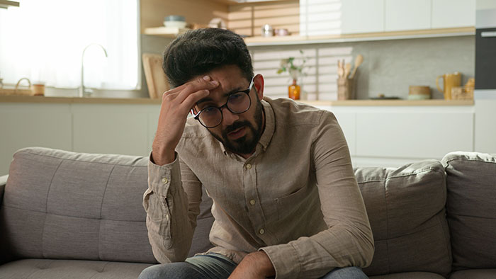 Man in glasses sitting on a couch, appearing frustrated, in a modern living room setting. Man in glasses sitting on a couch, appearing frustrated, in a modern living room setting.