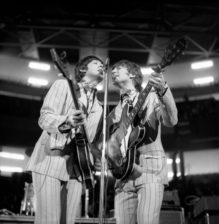 Two members of an iconic duo from a famous band, standing in suits with musical instruments in the background.