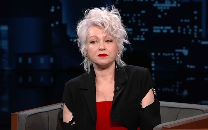 Cyndi Lauper with short, tousled white hair and red lipstick, in a black outfit on a TV show set.