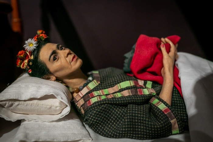 Hyper-realistic sculpture by Ruben Orozco Loza of a woman with flowers in her hair lying on a bed holding a red cloth.