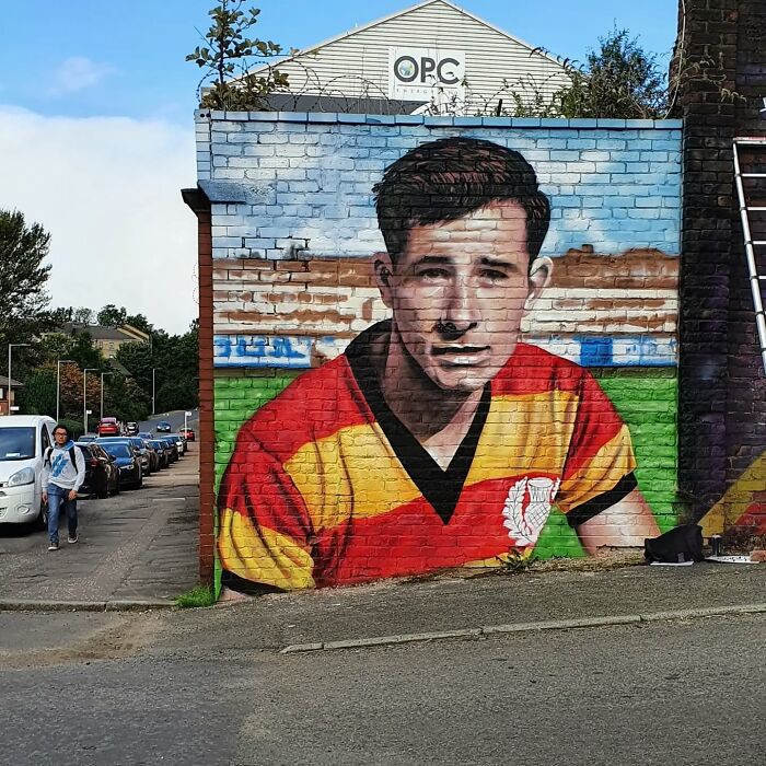 Mural of a footballer in a red and yellow jersey on a Glasgow wall, capturing local art essence.
