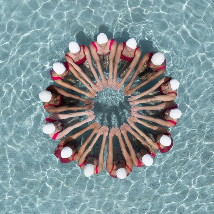 Aerial conceptual photography showing a circle of swimmers in red swimsuits and white caps in a pool.