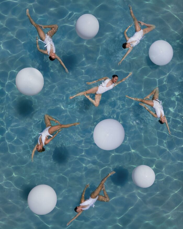 Aerial conceptual photography of synchronized swimmers in a pool with large white balls.