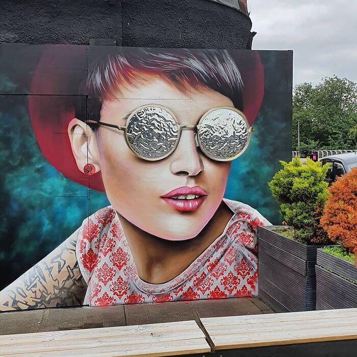 Vibrant mural of a woman in reflective glasses on a Glasgow wall, blending urban art with natural surroundings.