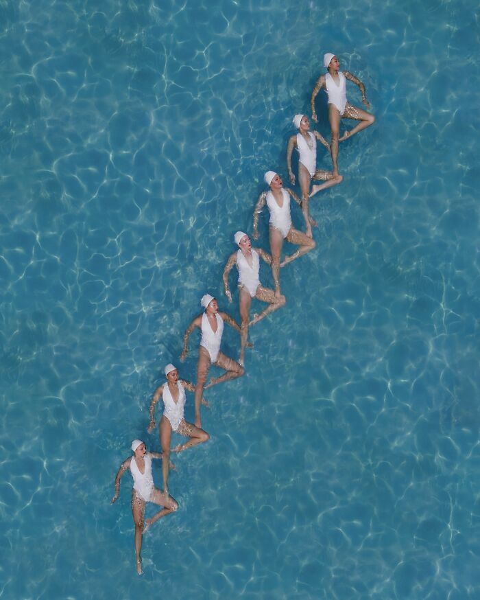 Aerial view of seven synchronized swimmers in white swimsuits forming a diagonal line in a pool.