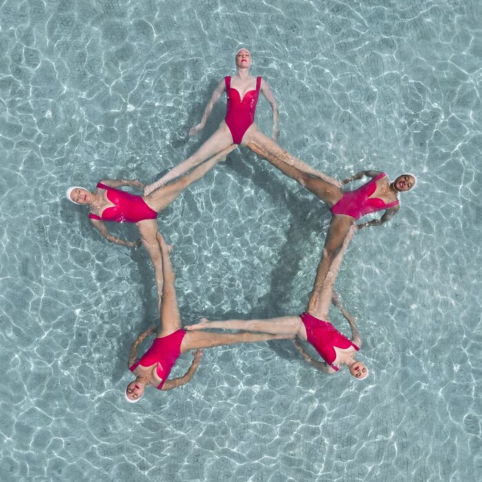 Aerial view of five synchronized swimmers in red suits forming a star shape in clear blue water.
