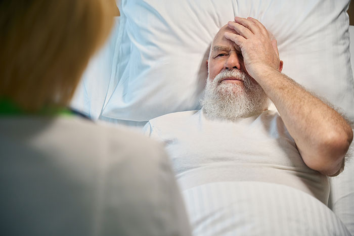 Elderly man in bed holding his head, appearing distressed during a medical consultation.