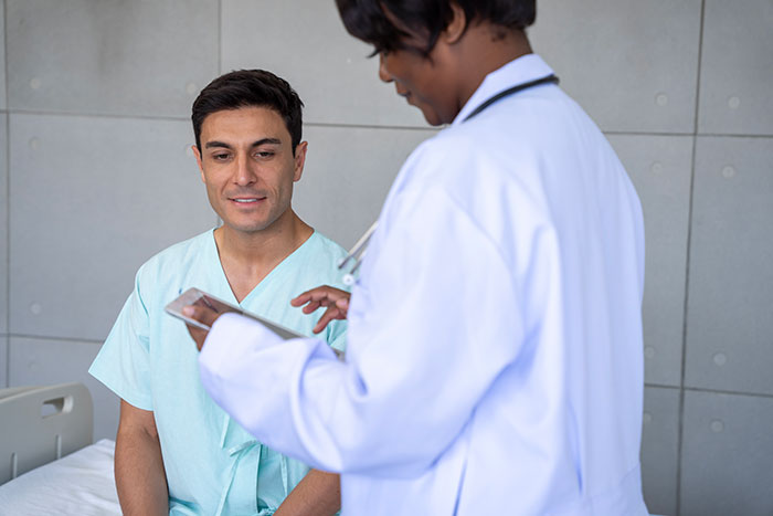 Doctor discussing medical diagnosis with patient in hospital gown, checking notes on a tablet.