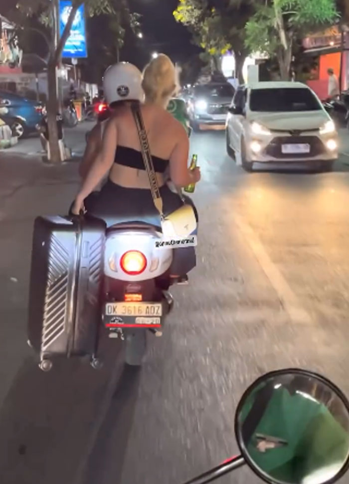 Tourist riding scooter at night holding a large suitcase and a drink, showing entitled and rude tourist behavior.