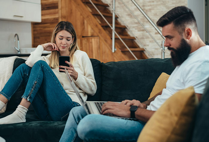 Couple sitting on a couch, woman looking at her phone, man using a laptop, discussing wedding planning frustration.