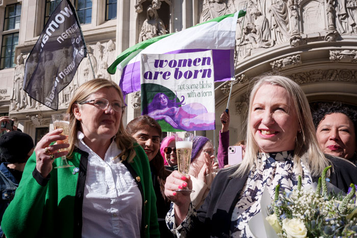 Women holding a banner and drinks outside a court, celebrating a ruling related to trans rights.