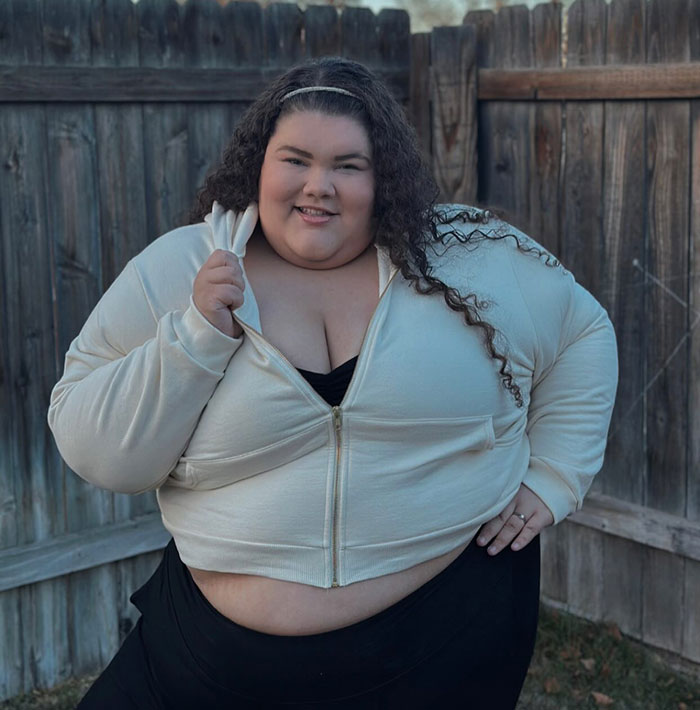 Plus-sized woman wearing a light hoodie outdoors, against a wooden fence, expressing a confident demeanor.