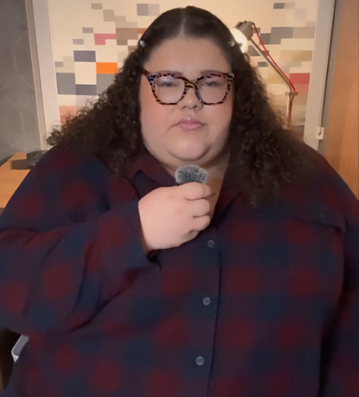 Plus-sized passenger with glasses holding a microphone, expressing concern in an indoor setting.