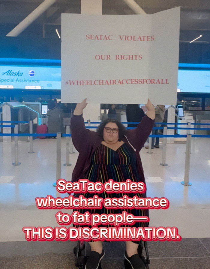 Plus-sized passenger at airport holding sign about wheelchair access rights and discrimination.