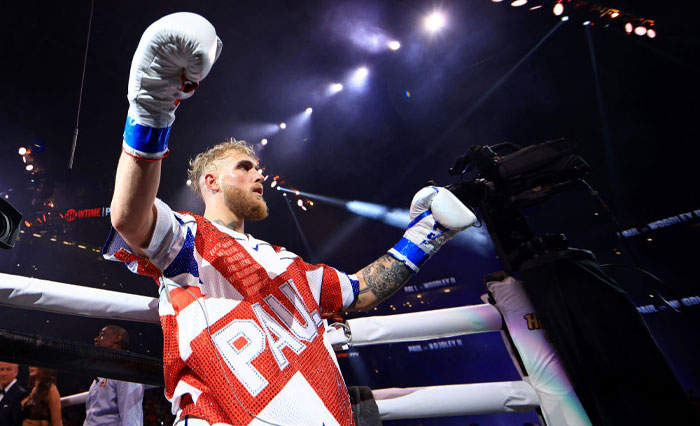 Jake Paul in red and white robe with gloves raised in a lit-up ring, showcasing YouTube fame and success beyond the platform.