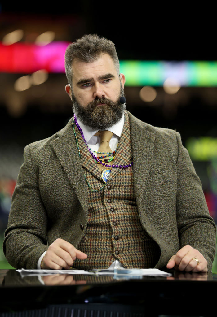 Jason Kelce in a tweed suit and tie with a microphone headset, focusing on building his fortune.