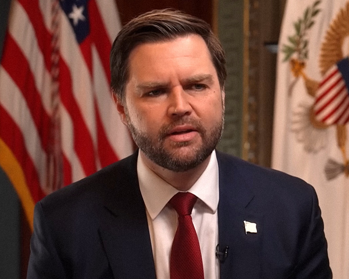 Man in a suit and red tie speaking, U.S. flags in the background.