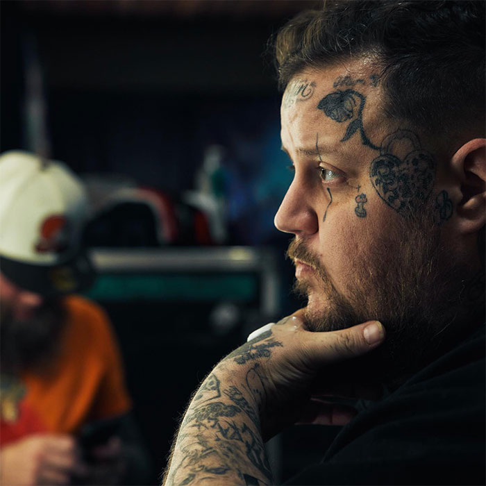 Jelly Roll contemplating in a dimly lit room, showcasing face tattoos and artistic expression, highlighting his net worth journey.