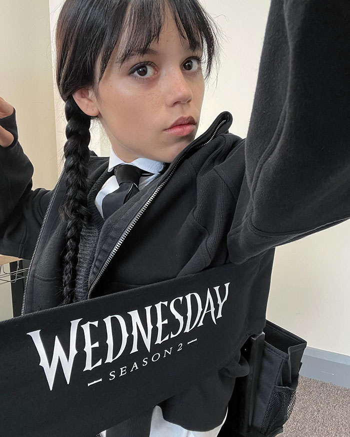 Young woman with a braided hairstyle, wearing a black jacket, promoting Wednesday Season 2.