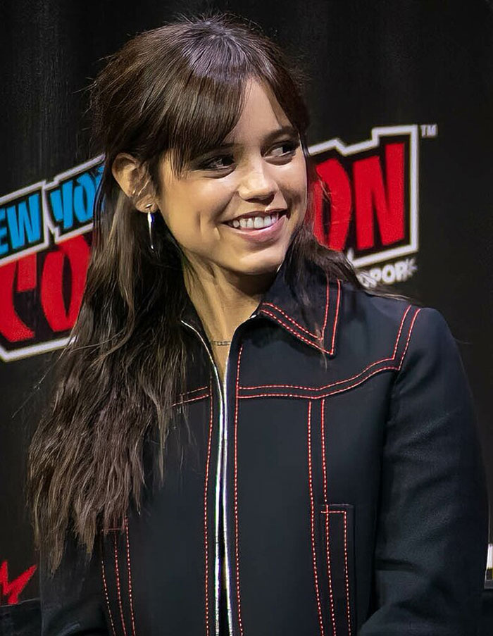 Young woman at a comic convention, wearing a black jacket with red stitching, smiling at the audience.