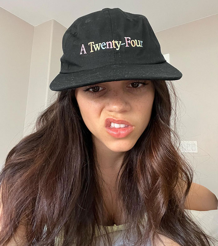 Person wearing a black cap inside a room, showcasing a new expression and hairstyle.